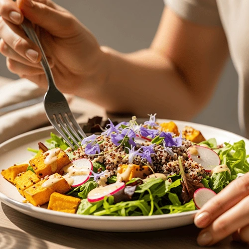 Un primer plano de una persona comiendo de un plato con una ensalada colorida y saludable, incluyendo quinoa y batatas, en un ambiente luminoso. La imagen representa la alimentación consciente y el disfrute de la comida sana.