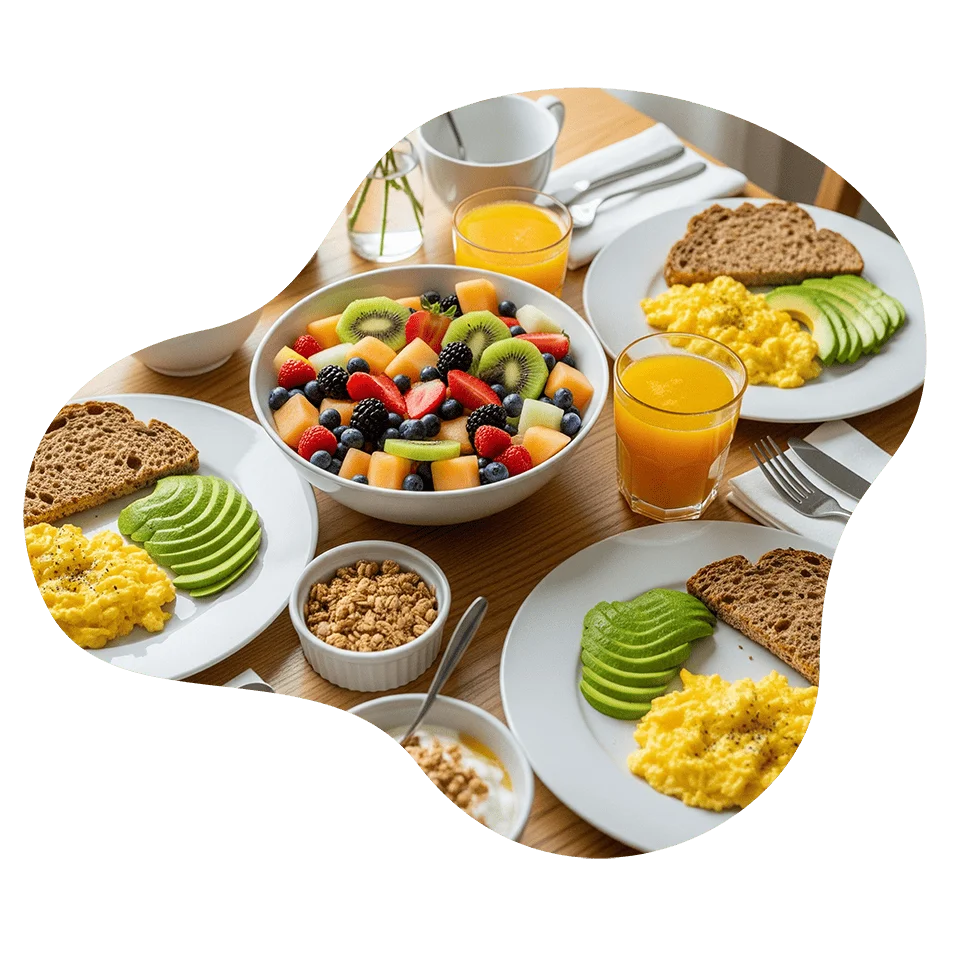 Mesa de desayuno saludable. Aparece un bol de frutas variadas en medio y cuatro desayunos compuestos por tostadas integrales, aguacate, huevos revueltos, cereales y zumo de naranja.